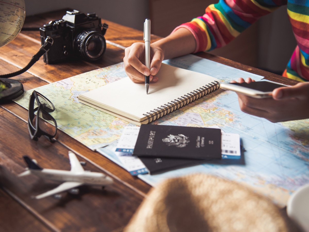 Person writing in a notebook with a map, passports, camera and phone on desk showing how to plan an itinerary
