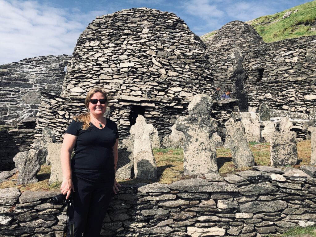 The author and travelpreneur, Erica Forrest holding a camera in front of the dry stacked monk dwellings on the island of Skellig Michael in Ireland.