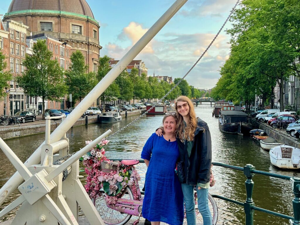 The author, Erica Forrest with her daughter on a canal bridge in Amsterdam with a pink floral decorated bike, canal boats, and old buildings in the background.